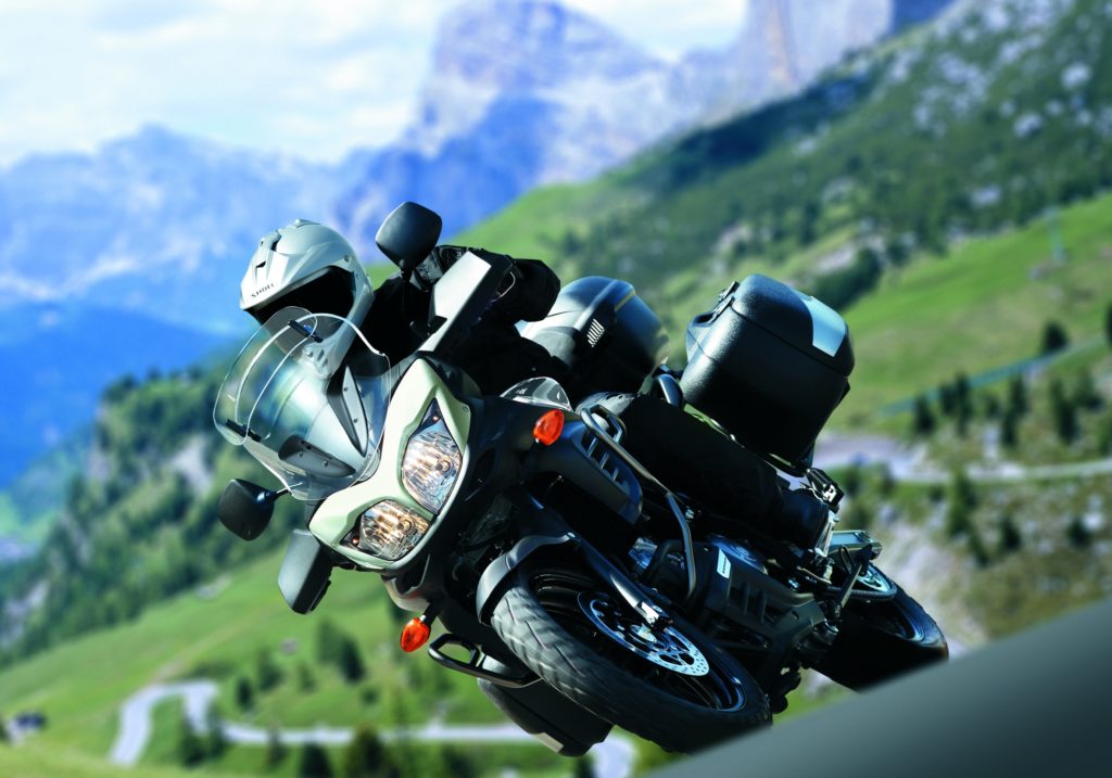 Ein Motorradfahrer auf einer Suzuki fährt dynamisch durch eine malerische Berglandschaft mit grünen Wiesen und hohen Bergen im Hintergrund.
