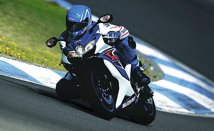 Ein Fahrer auf einer Suzuki-Motorradkurvenfahrt auf einer Rennstrecke, umgeben von grünem Gras und blühenden Blumen.
