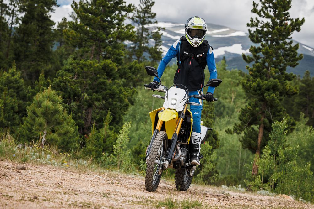 Motorradfahrer in Schutzkleidung fährt auf einem gelben Offroad-Motorrad durch eine bewaldete Landschaft mit Bergen im Hintergrund.