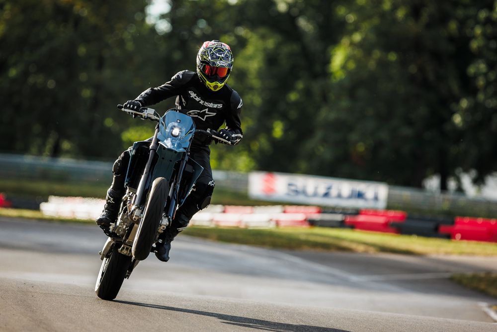 Motorradfahrer in Schutzkleidung vollführt einen Wheelie auf einer Rennstrecke mit Suzuki-Werbung im Hintergrund.