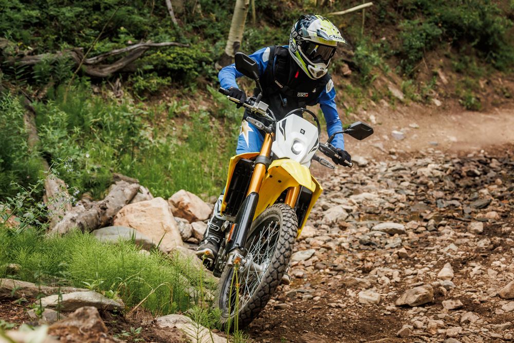 Ein Motorradfahrer in Schutzkleidung fährt auf einem unebenen, steinigen Weg durch eine grüne Landschaft.