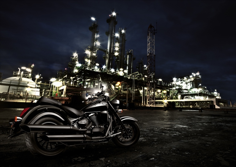 Ein schwarzes Motorrad steht vor einer beleuchteten Industrieanlage bei Nacht, was eine kraftvolle und dynamische Atmosphäre schafft.